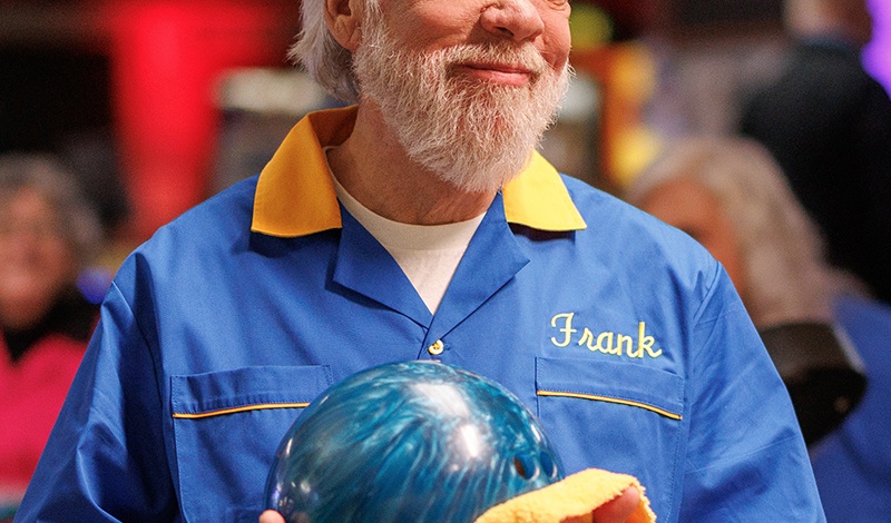 Smiling older man in a blue bowling shirt holds a bowling ball and towel inside a lively bowling alley.