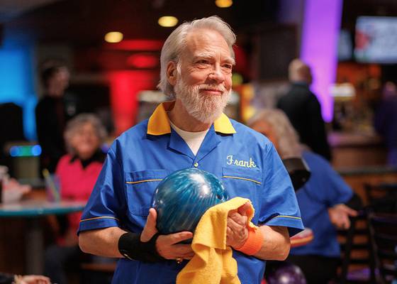Smiling older man in a blue bowling shirt holds a bowling ball and towel inside a lively bowling alley.