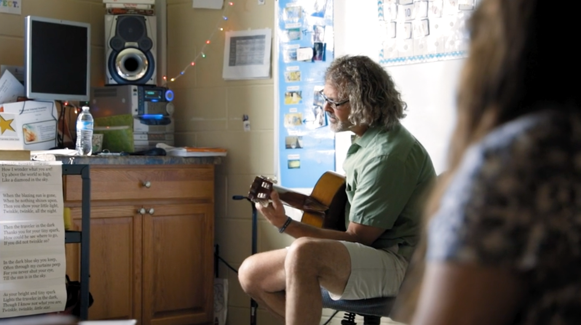 Still from video of man playing guitar in background with out of focus woman in foreground.